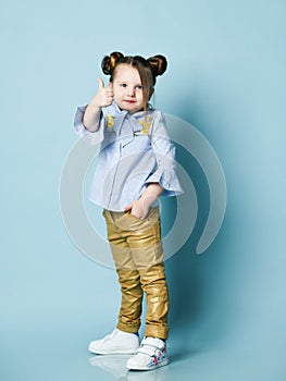 Calm and contented kid girl with funny buns and in blue shirt and gold leather pants is showing thumbs-up sign gesture
