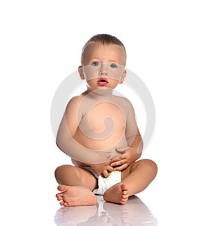 Calm and curious baby boy toddler in diaper sits on the floor, touches his stomach and looks at camera