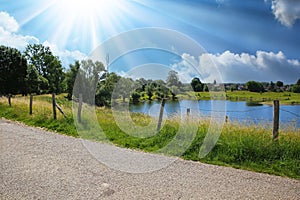 Calm cycling track in beautiful rural dutch landscape, small lake on farm land, green meadows, blue summer sky with sun rays -