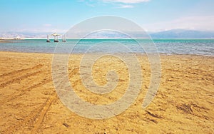 Calm day at Ein Bokek Dead Sea beach, blue green water, sun shade shelter near, sun shines on sandy beach shore