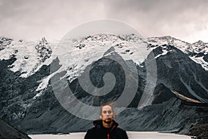 calm disheveled young caucasian man bundled up in black coat in front of huge snowy big rocky mountains over calm lake