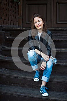 A calm girl in sneakers, jeans and a leather jacket sits on wood