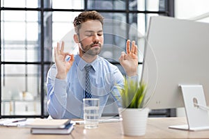 Calm handsome businessman meditate keeping mudra listening to relaxing music at office desk, meditation.