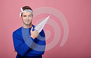 Calm happy guy in bandana in dark blue sweater holds white paper airplane in his hand and shows at left