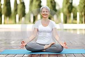 Calm happy senior woman sitting in lotus pose on mat during morning meditation in park