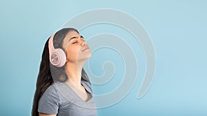 Calm Indian teen girl wearing wireless headphones, listening to music with closed eyes on blue background, copy space