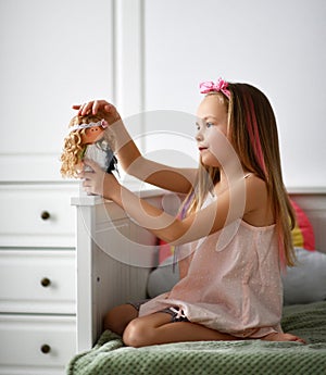 Joyful kid girl in home clothing dress and headband with bow plays with her favorite doll in her bed at home