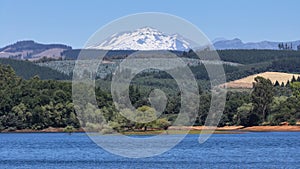 Calm lake with a green forest and the snowy Nevados de Chillan mountain in the background