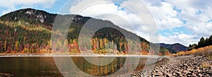 Calm lake with low water - round stones at shore visible, autumn coloured coniferous trees on other side, blue sky above