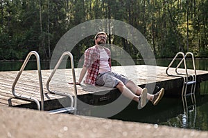 Calm man sits on the edge of a wooden pier and looks up thoughtfully, against the background of a calm forest lake