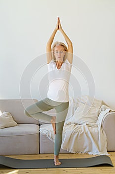 Calm mature woman in sports wear standing in Tree pose on yoga mat at home