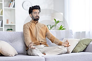 Calm middle eastern man sitting on pillowed sofa in siddhasana position and using hands mudra with closed eyes. Calm guy