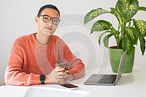 Calm millennial latin lady enjoy work and cup of coffee at table with computer