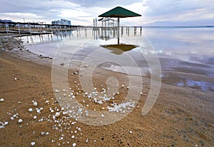 Calm morning at Ein Bokek Dead Sea beach, clouds reflection over water surface near sun shade shelter, sand covered with
