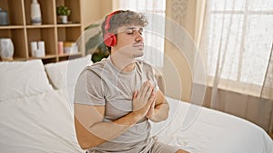 Calm morning indoors, attractive young hispanic man finds balance, relaxing on bed in pjs, meditating to music through headphones