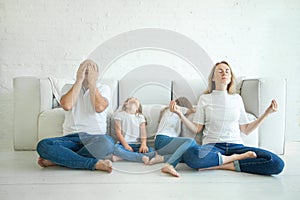 Calm mother with closed eyes meditating in lotus pose on floor, stressed father and two tired children in living room at home