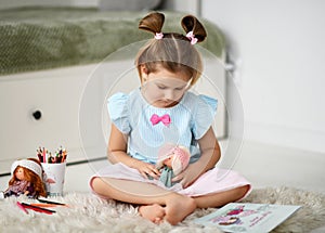 Calm and peaceful kid girl sits on soft carpet at home playing stories with her dolls and drawing with colorful pencils