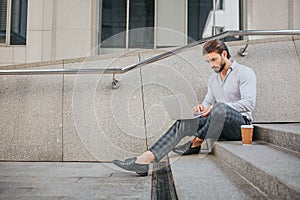 Calm and peaceful young man sits on steps and works. He looks at laptop screen. There are plastic cup of coffee on steps