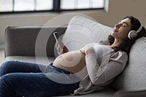 Calm Pregnant Lady In Wireless Headphones Listening Music On Smartphone At Home