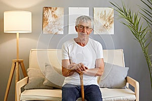 Calm relaxed man pensioner wearing white T-shirt and jeans sitting on sofa in living room holding walking cane looking at camera