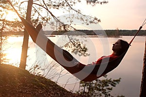 Calm relaxed woman lying in hammock in forest admiring lake nature at sunset.