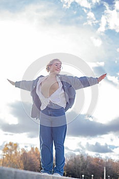 Calm satisfied spiritual woman in jeans outfit stand on border with raised arms like in Titanic against sunny cloudy sky