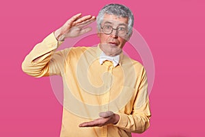 Calm senior shows large or big object, stands against pink wall, dressed in stylish yellow shirt and white bowltie, has serious