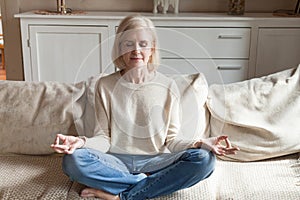 Calm senior woman practicing yoga meditating on couch