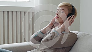 Calm serene young adult woman relaxing on comfortable sofa eyes closed wear headphones enjoy listen favorite cool music