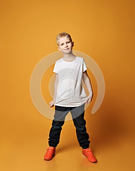 Calm, smart young schoolboy kid in blue jeans and sneakers stands showing his white blank t-shirt