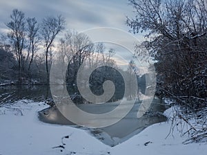 Calm snowy winter scene with river, snow covered riverbanks and naked trees