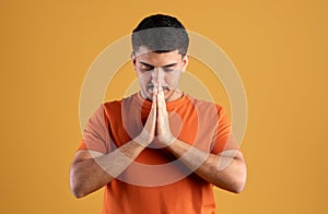 Calm spiritual brazilian man praying with closed eyes, peaceful male with joining hands meditating on yellow background