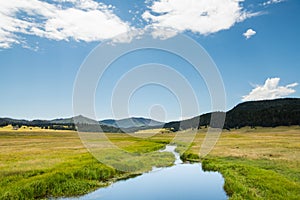 A calm stream reflects a beautiful blue sky as it curves through lush green meadows in the mountains of northern New Mexico