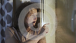 Calm tousled young woman sits by window. Girl draws with her finger on glass.