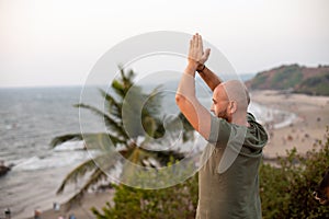 Calm vacationist folding palms together and lifting arms over head standing on hill near plants and looking at shore.