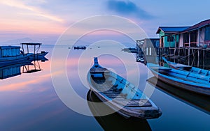 Calm waters reflect a sublime sunset at a peaceful fishing village, with traditional boats moored beside rustic stilt