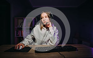 Calm woman in a headset sitting at home at the computer and playing online video games at night, streaming, looking away at the
