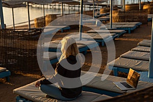 A calm woman sits on a sun lounger near the sea, gets distracted from working at the computer, meditates and removes