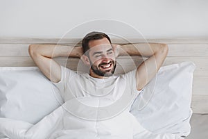 Calm young handsome bearded man lying in bed with white sheet pillow blanket in bedroom at home. Smiling beauty male