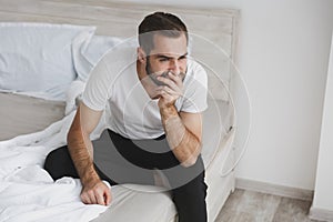 Calm young handsome bearded man sitting in bed with white sheet pillow blanket in bedroom at home. Sad upset male