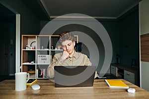 Calm young male freelancer in shirt working on laptop in cozy bedroom at home. Guy in the wireless headphones works remotely over