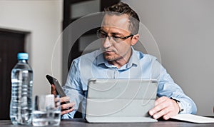 Calm young man in shirt and trousers sitting in the office with a smartphone in his hand and looking at it