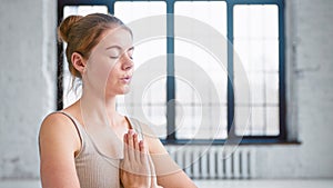 Calm young woman with hair bun in beige top does yoga breathing exercises holding hands in namaste mudra in studio