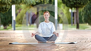 Calm young woman sitting in lotus pose on mat during morning meditation in park