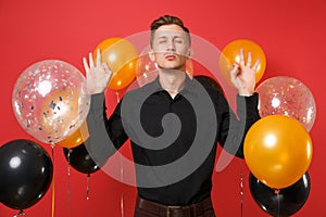 Calmed young man in black classic shirt meditate spreading hands on bright red background air balloons. St. Valentine`s