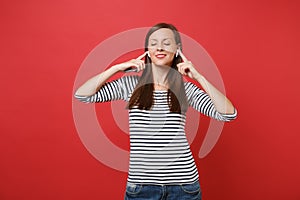 Calmed young woman in striped clothes with wireless earphones holding mobile phone, listening music isolated on bright