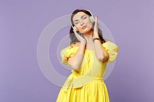 Calmed young woman in yellow dress keeping eyes closed, listen music, putting hands on headphones isolated on pastel