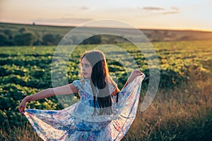 Calmy beautiful brunette teen girl with loose hair atanding among the mealow, holding flying dress with flowers print in