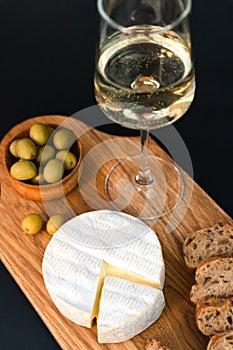 Camembert cheese, crispy baguette, olives and wineglass of white wine on a dark background. Close-up. Selective focus