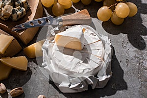 Camembert near grana padano, dorblu, knife on wooden board with grapes and nuts on grey background.